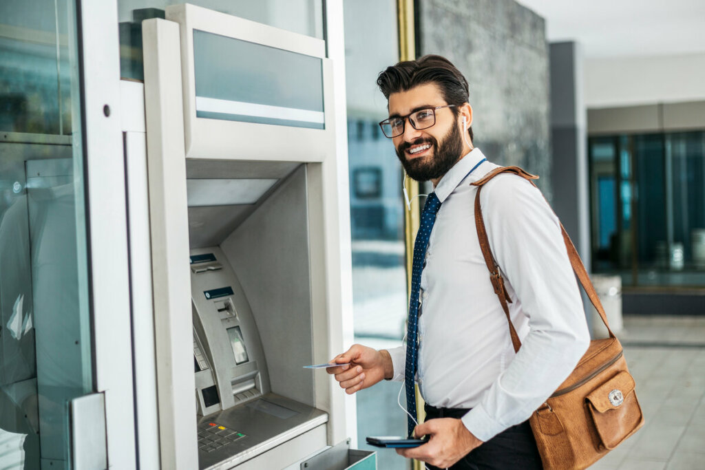 Homem vestindo roupa social e com uma bolsa bege em frente a um caixa eletrônico