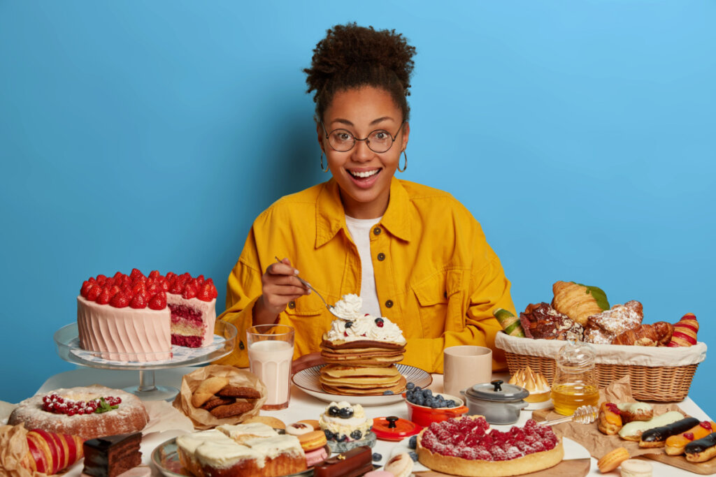 Mulher com blusa amarela de frente para uma mesa cheia de alimentos gordurosos e cheios de açúcar, tais como bolo, panqueca, croissant, tortas, chocolates e mais