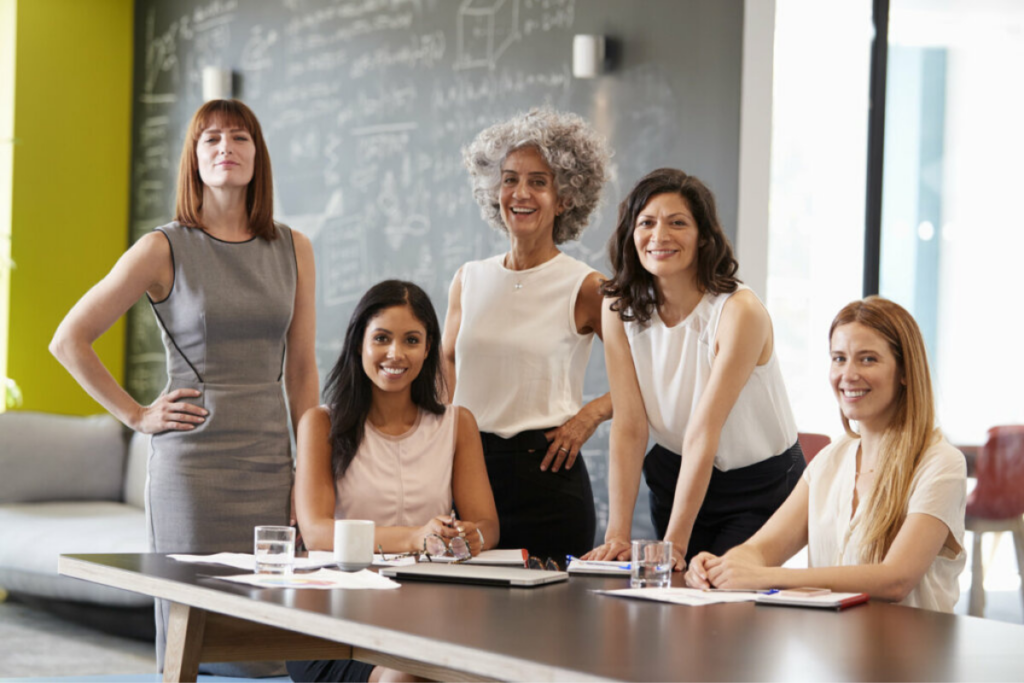 Cinco mulheres de diferentes faixas etárias sorrindo para a câmera em uma reunião de trabalho