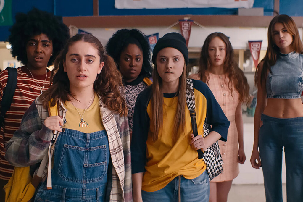 Grupo de seis meninas adolescentes, usando roupas coloridas e de estilos variados. Elas olhando sérias e preocupadas para a mesma direção, e estão em um corredor de um colégio tipicamente americano