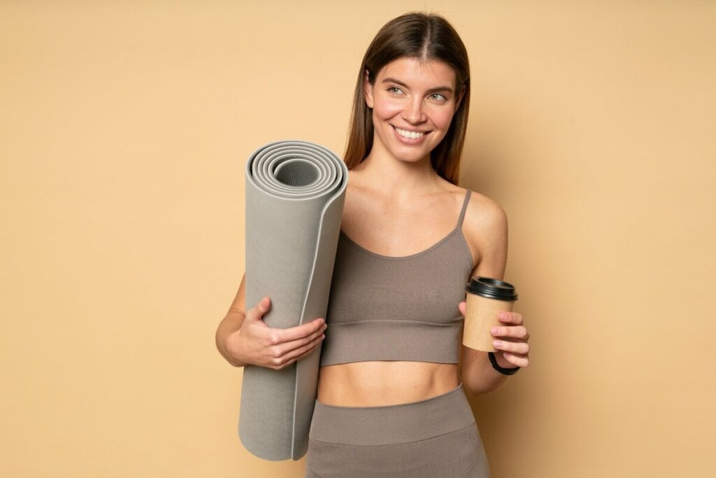 Mulher com roupa de academia sorrindo segurando um copo de café e um tapete e ioga