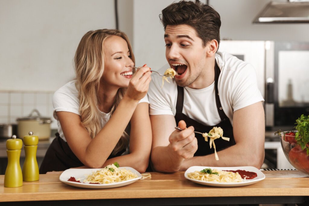 Mulher loira colocando garfo com macarrão na boca de homem. Eles estão na cozinha