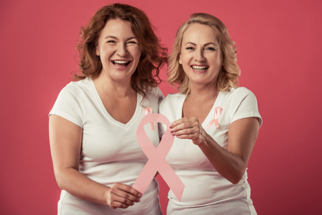 Duas mulheres sorrindo e segurando laço rosa em referência ao câncer de mama