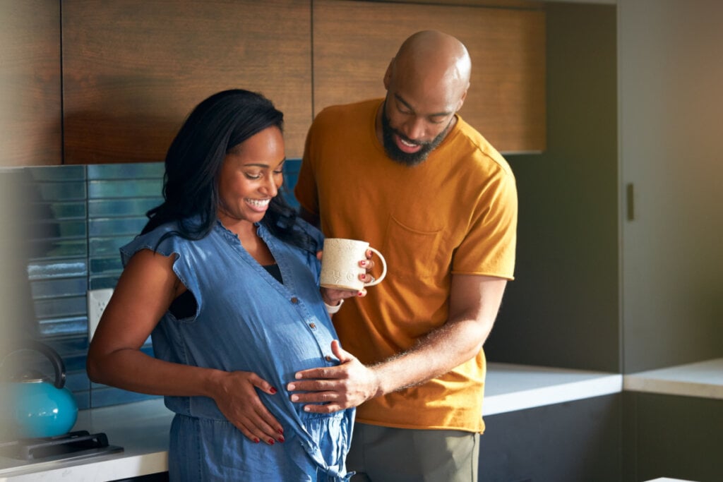 Mulher segurando caneca e com a mão na barriga. Homem ao lado dela também toca a barriga