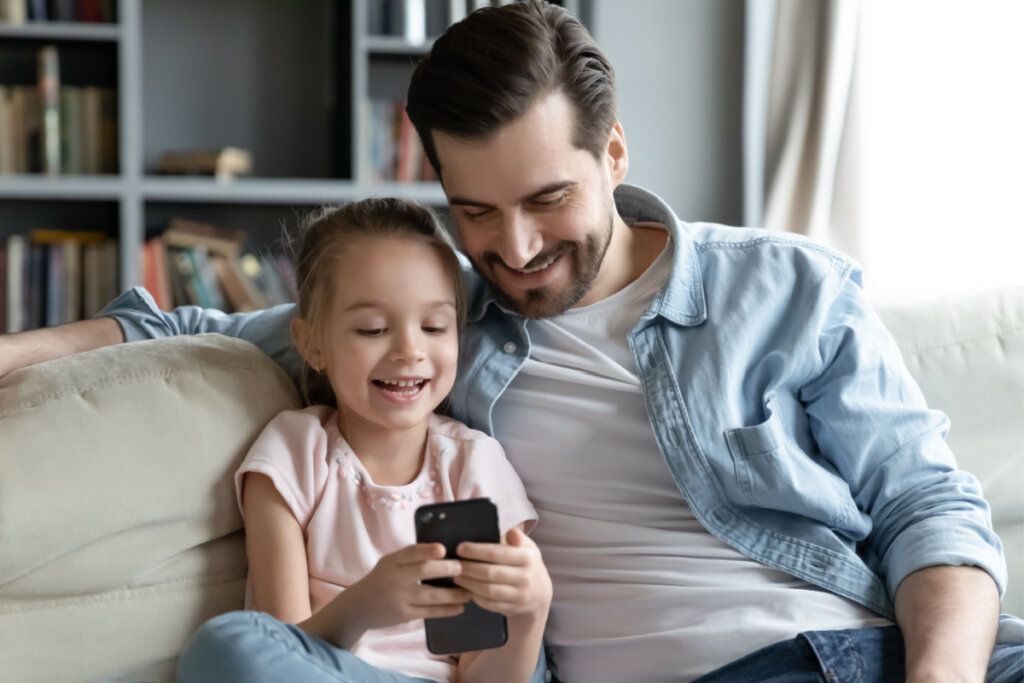 Pai e filha pequena sentados no sofá. Ela mexe em celular preto