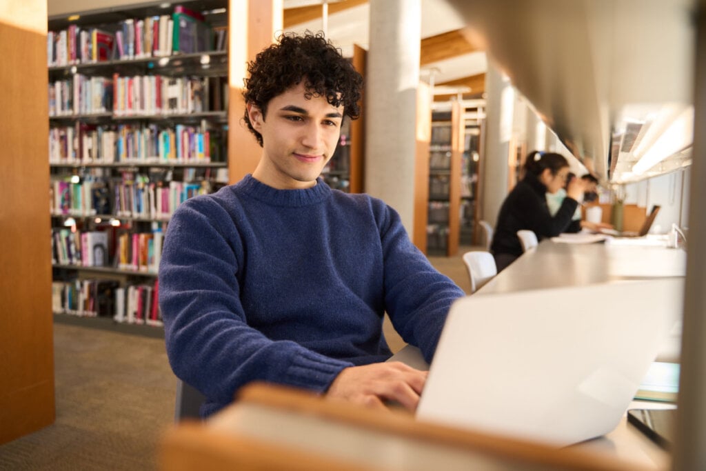 Homem jovem, de blusa azul, estudando em um laptob na biblioteca de uma universidade