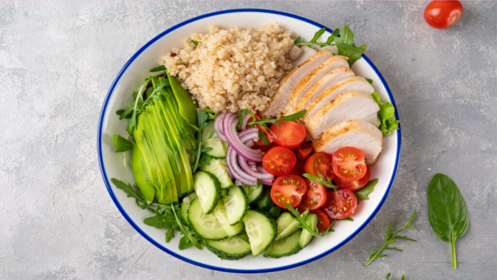 Tigela branca com borda azul e salada de quinoa, frango, tomate, pepino, cebola-roxa, abacate e rúcula