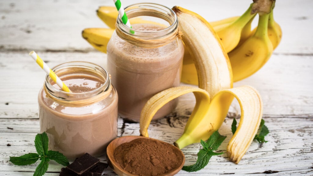 Smoothie de banana com whey sabor chocolate em dois copos separados ao lado de bananas e whey