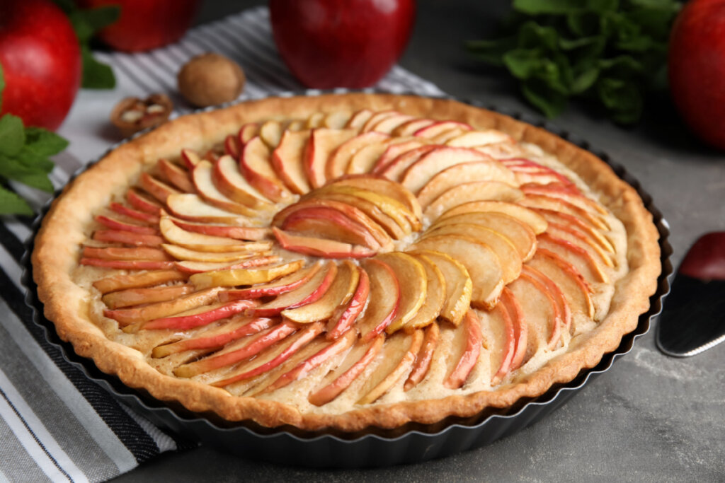 Torta de maçã com fatias da fruta cobrindo a massa, em uma mesa com maçãs ao fundo