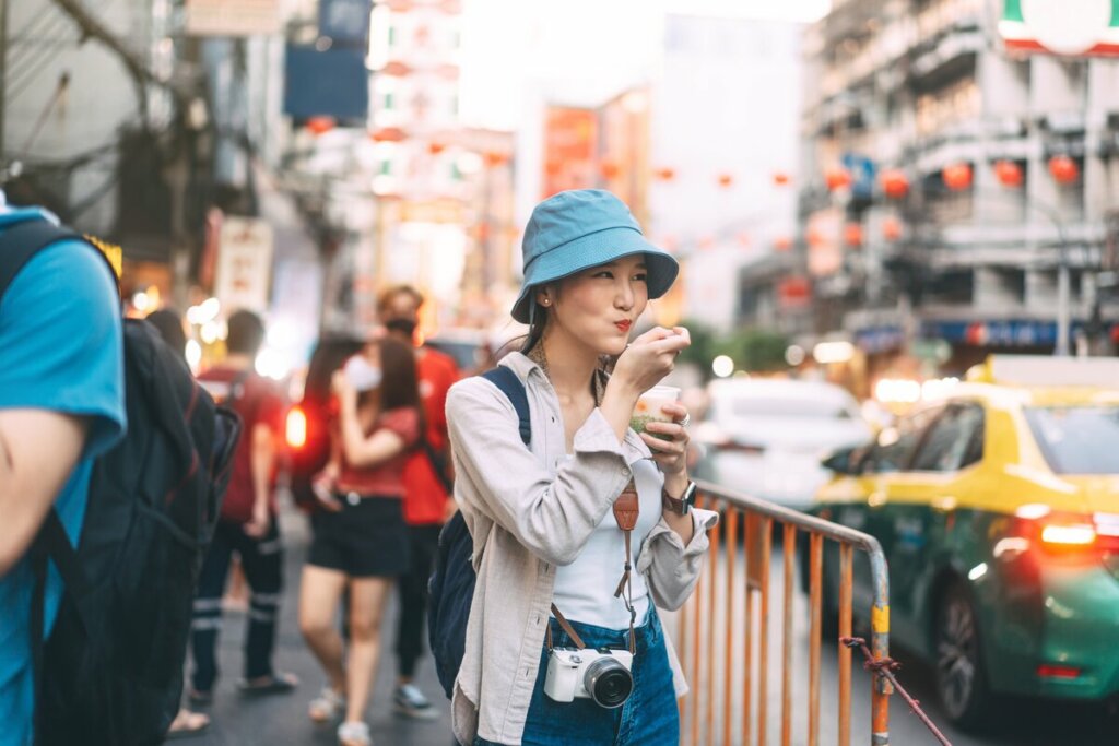 Mulher usando chapéu e com mochila nas costas comendo na rua