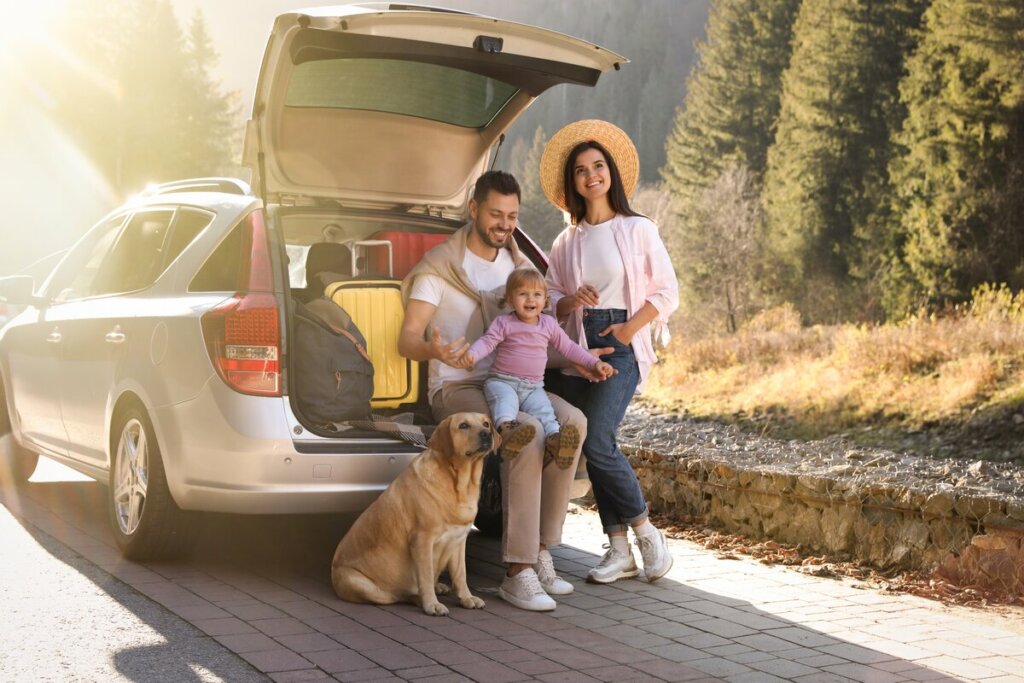 Homem, mulher, bebê e um cachorro em frente ao porta-malas de um carro sentados