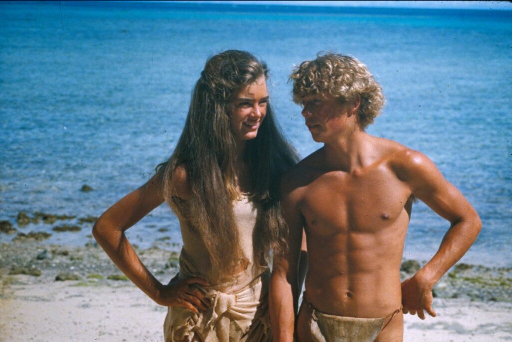 Emmeline e Richard do filme 'A Lagoa Azul' parados lado a lado em uma praia 