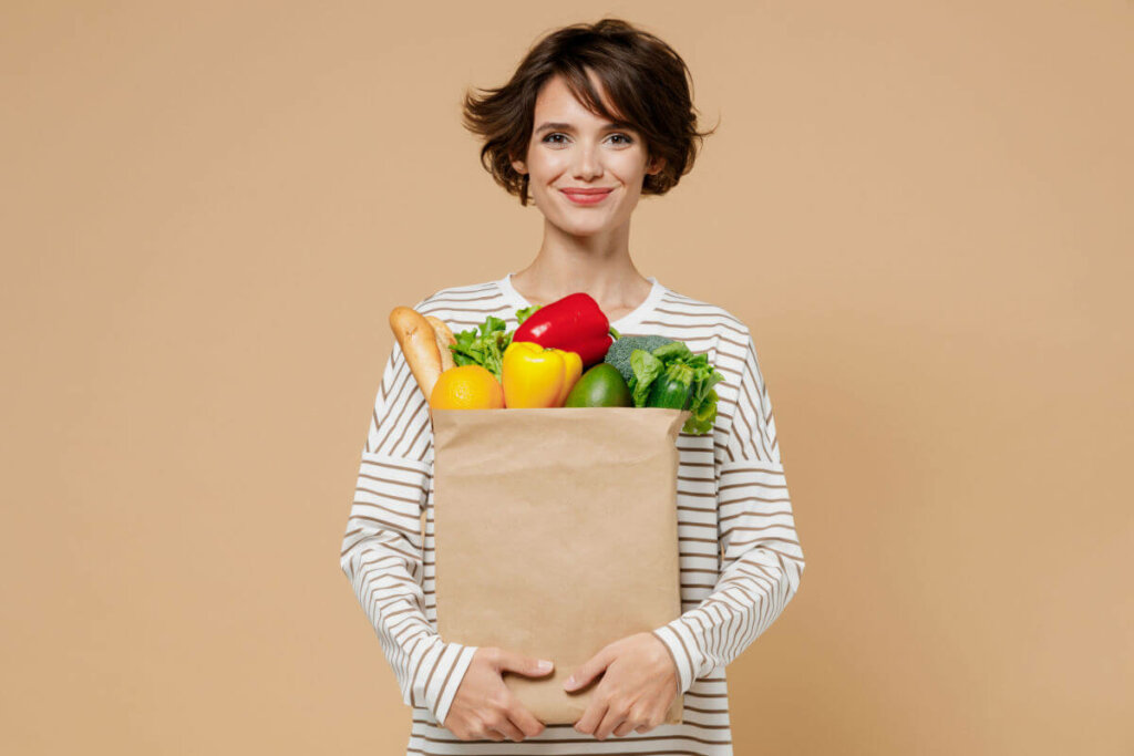 Mulher sorrindo segurando uma sacola com vegetais em frente a um fundo marrom