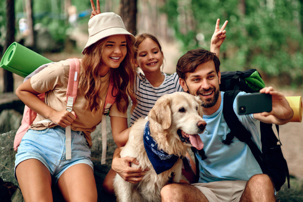 "Pai, mãe e filha, com cachorro Golden Retriever, tirando selfie com mochilas nas costas, indicando viagem