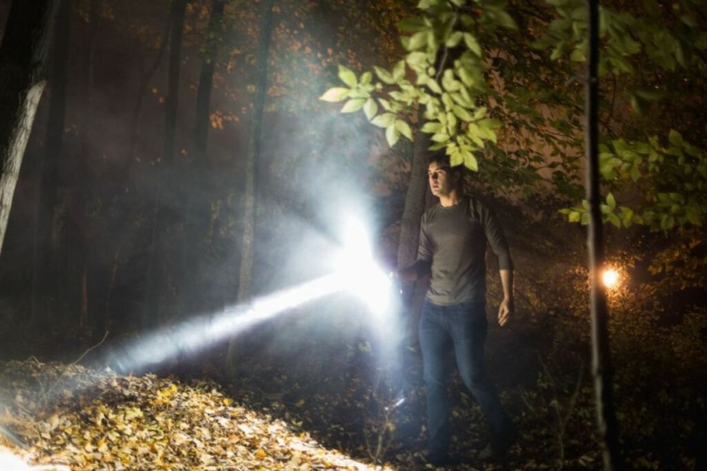 Cena do filme 'Mercy: Golpe de Misericórdia' com um homem segurando uma lanterna em uma floresta 