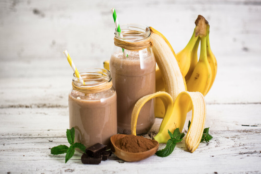 Dois copos com vitamina de banana e chocolate em cima de uma mesa de madeira com bananas e chocolate em pó