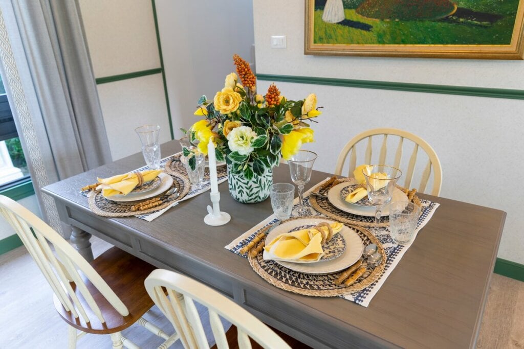 Mesa de jantar com vaso de flores amarelas, pratos brancos e cadeiras brancas