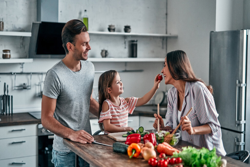 Pai, filha e mãe na cozinha preparando comida. Menina oferece um morango para a mãe