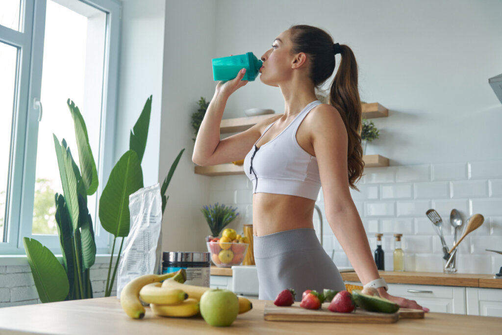 Mulher na cozinha com roupa de academia e bebendo em garrafa verde. Em bancada à frente dela tem diversas frutas