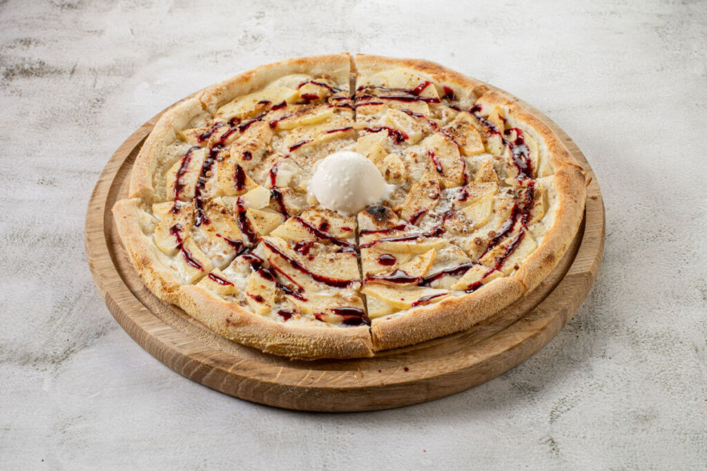 Pizza com pedaços de maçã, bola de sorvete e calda em cima de uma mesa branca