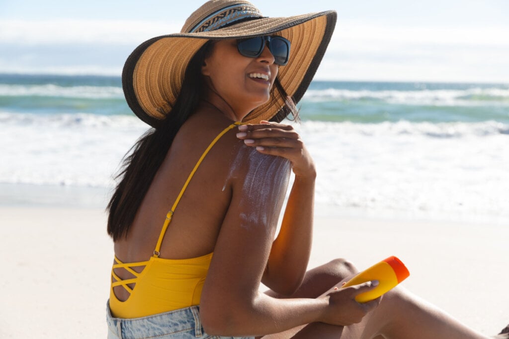 Mulher sentada na praia aplicando protetor solar no braço