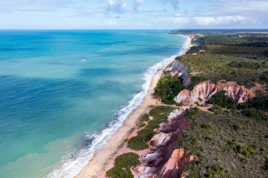 Vista aérea de praia com mar azul e morros com árvores e verde