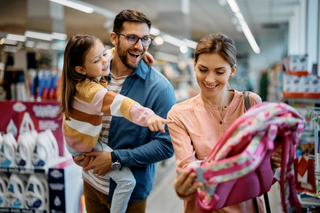 Menina feliz apontando para mochila enquanto compra material escolar com os pais