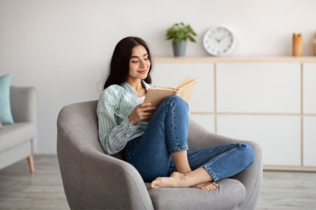 Mulher sentada em uma poltrona cinza e lendo um livro