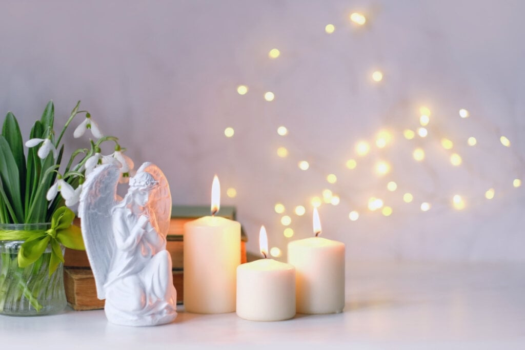 Anjo orante, velas, flores e livros sobre a mesa de perto, fundo de luz abstrata