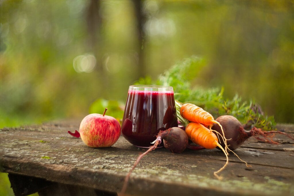 Copo de vidro com suco roxo com beterraba, cenoura e maçã ao lado