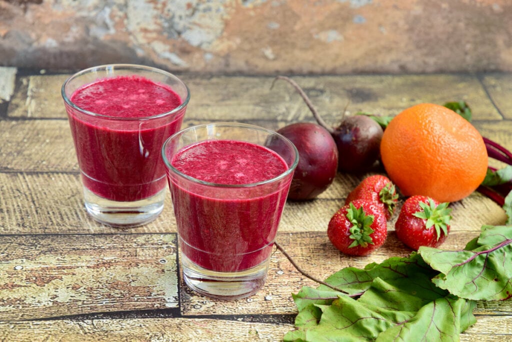 Dois copos com suco roxo em cima de uma mesa de madeira com beterraba, morango e laranja