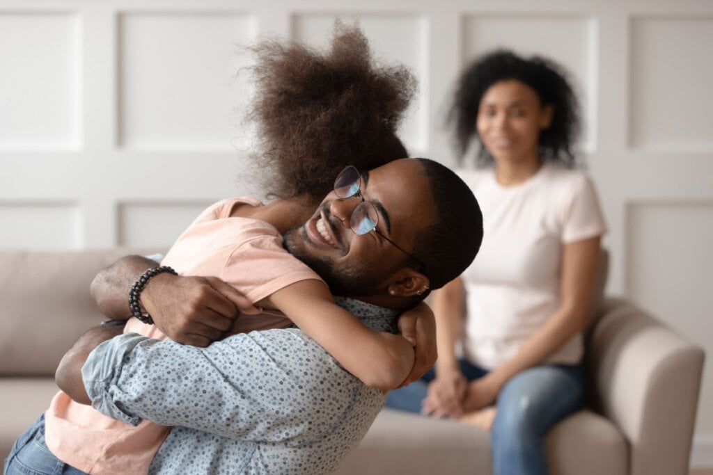 Pai abraçando filha e mãe ao fundo sentada em sofá