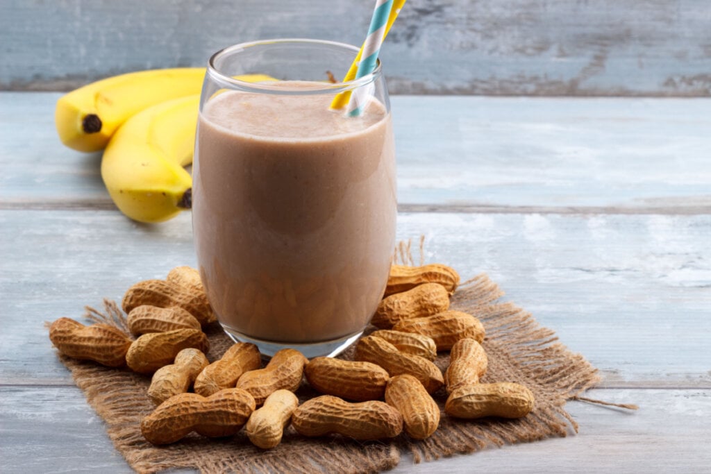 Copo de vidro com smoothie de banana com pasta de amendoim, com bananas e amendoins ao redor