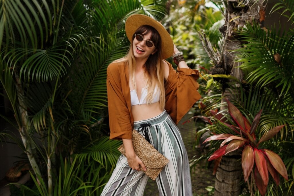 Mulher usando biquíni branco, chapéu, óculos, calça e um casaco posando para foto em frente a árvores