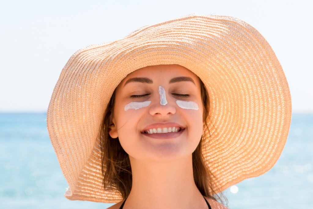 Mulher usando chapéu com dois riscos de protetor no rosto em uma praia 