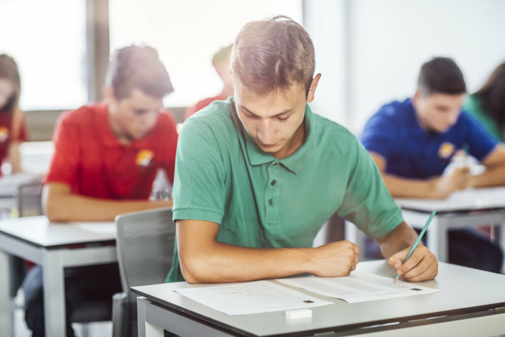Menino usando blusa verde sentado em uma sala de aula fazendo prova