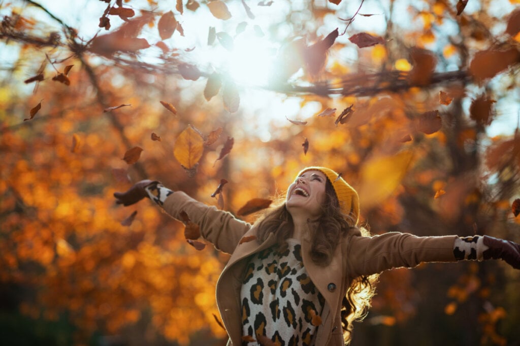 Imagem de uma mulher sorrindo com os braços para a cima em um cenário outonal