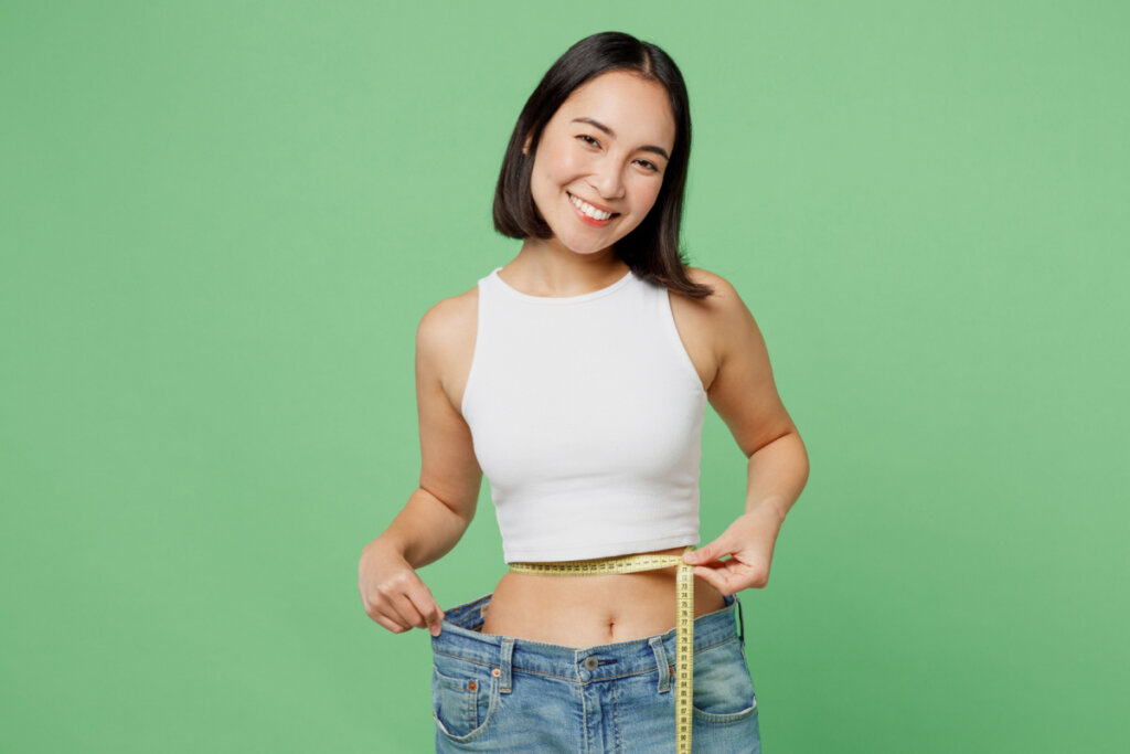 Mulher asiática sorrindo enquanto segura uma fita métrica na barriga e veste uma calça larga