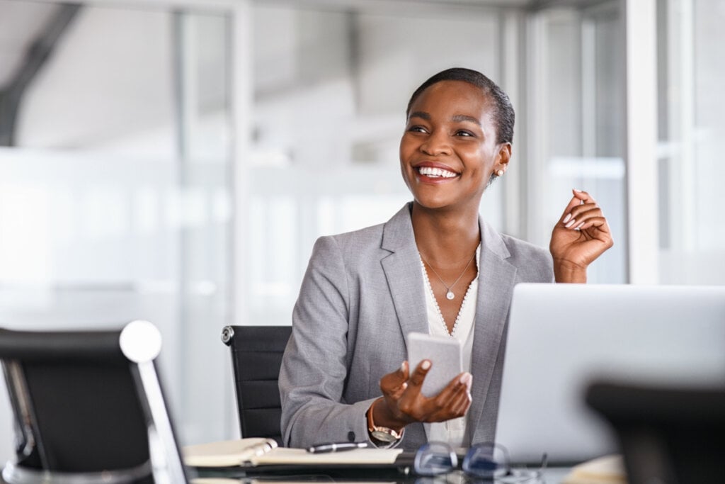 Mulher sorrindo com um celular na mão em uma sala de trabalho