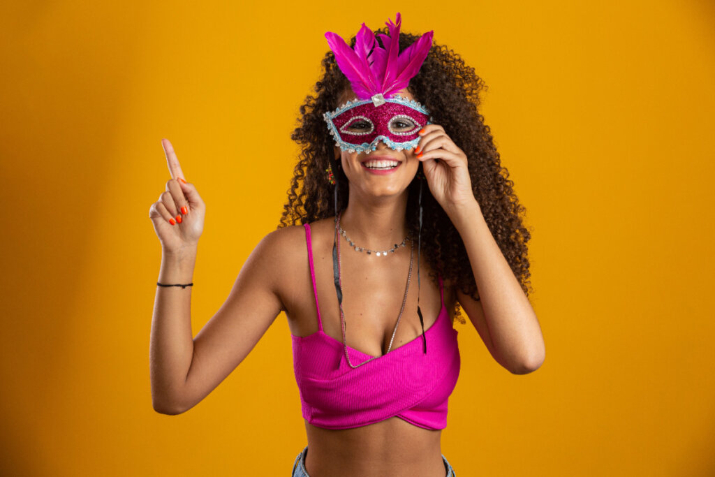 Mulher de cabelo cacheado, top cropped rosa e máscara rosa de Carnaval em fundo laranja