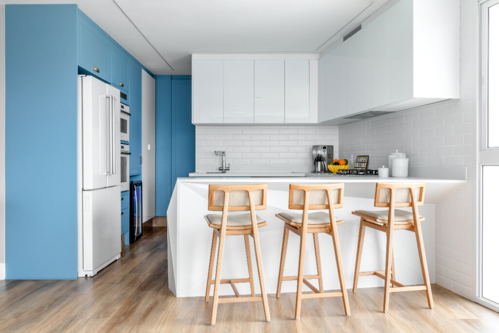 Cozinha com parede azul do lado esquerdo e uma geladeira branca; do lado direito, uma bancada com três banquetas e armários brancos
