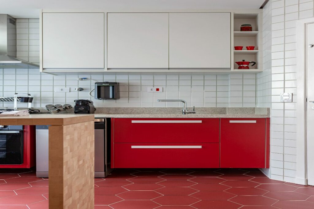 Cozinha com armário vermelho e piso vermelho em hexágonos; em cima, armários brancos e eletrodomésticos pretos e vermelhos