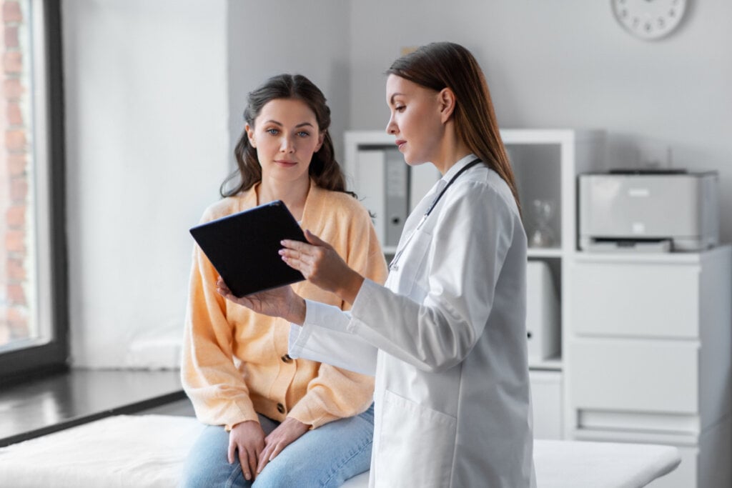 Médica mostrando algo em tablet para mulher sentada em maca