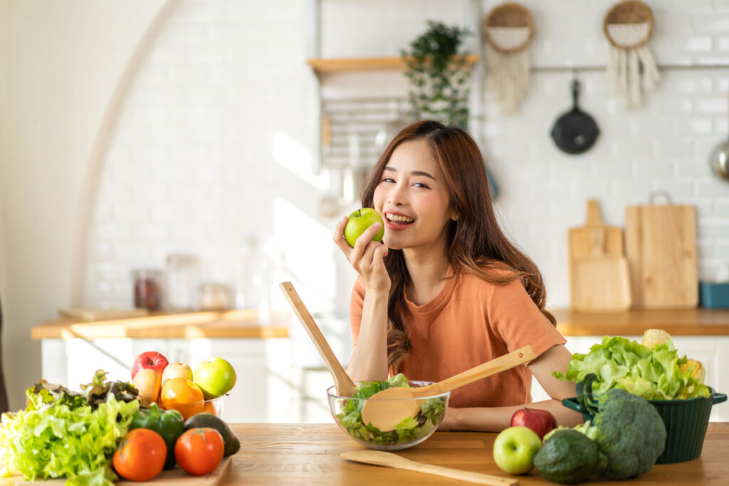 Mulher na cozinha comendo maçã e bancada com diversos vegetais
