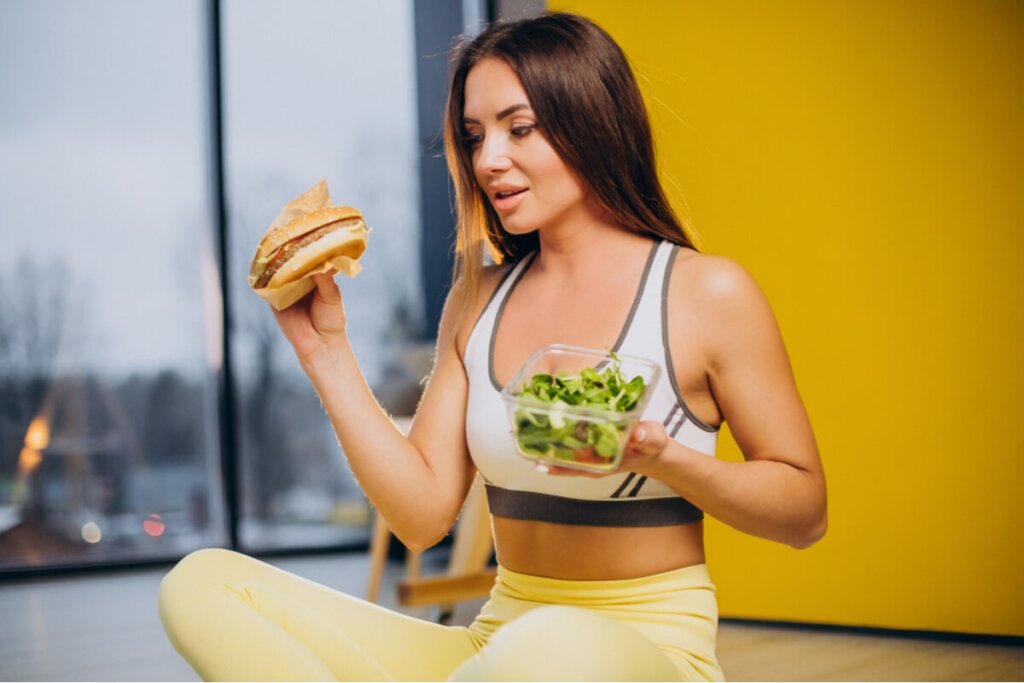 Mulher olhando para um hambúrguer e segurando pote de salada