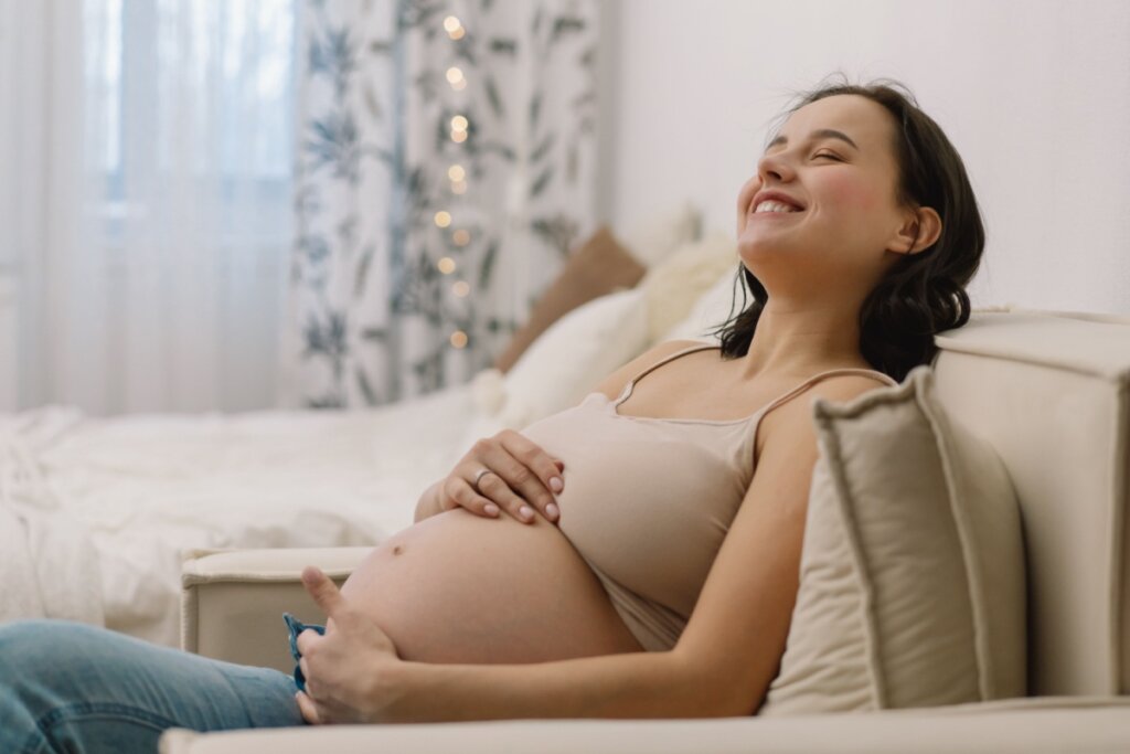 Mulher grávida sorrindo sentada no sofá com as mãos na barriga.