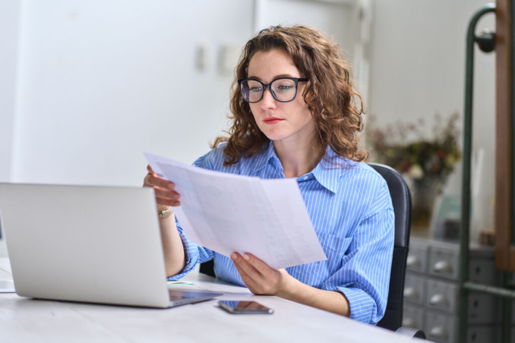 Mulher jovem segurando um papel sentada em uma mesa com notebook.