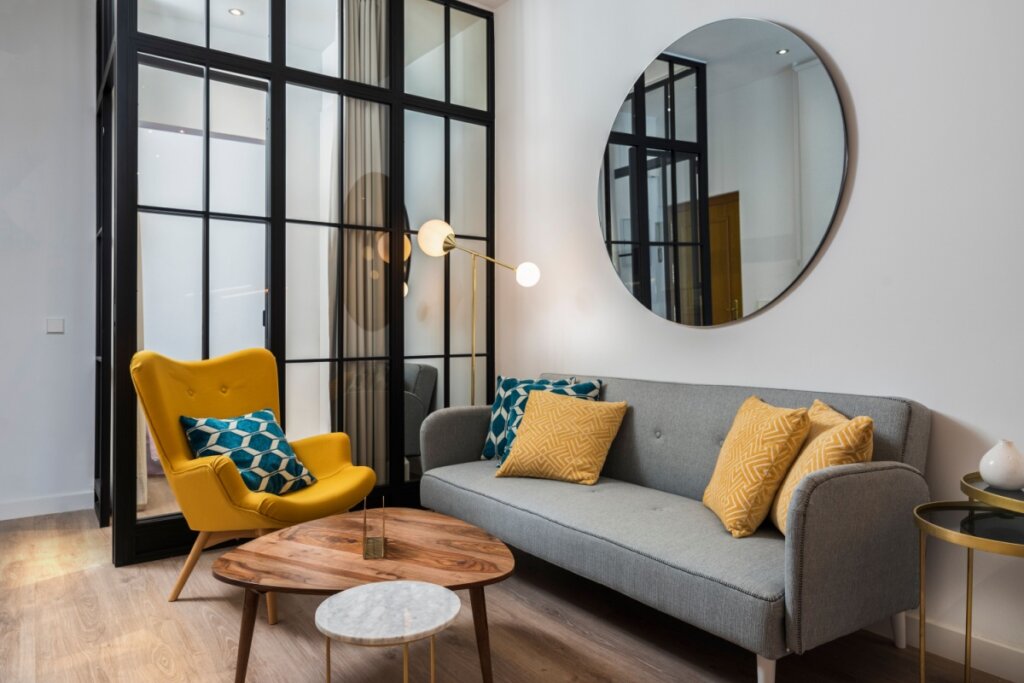 Sala de estar colorida e aconchegante com poltrona e sofá de design, além de um espelho decorativo redondo e parede de vidro.