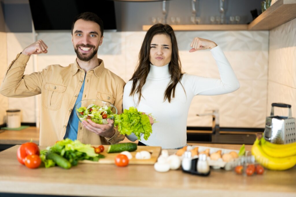 Casal na cozinha segurando alimentos saudáveis e mostrando músculo do braço