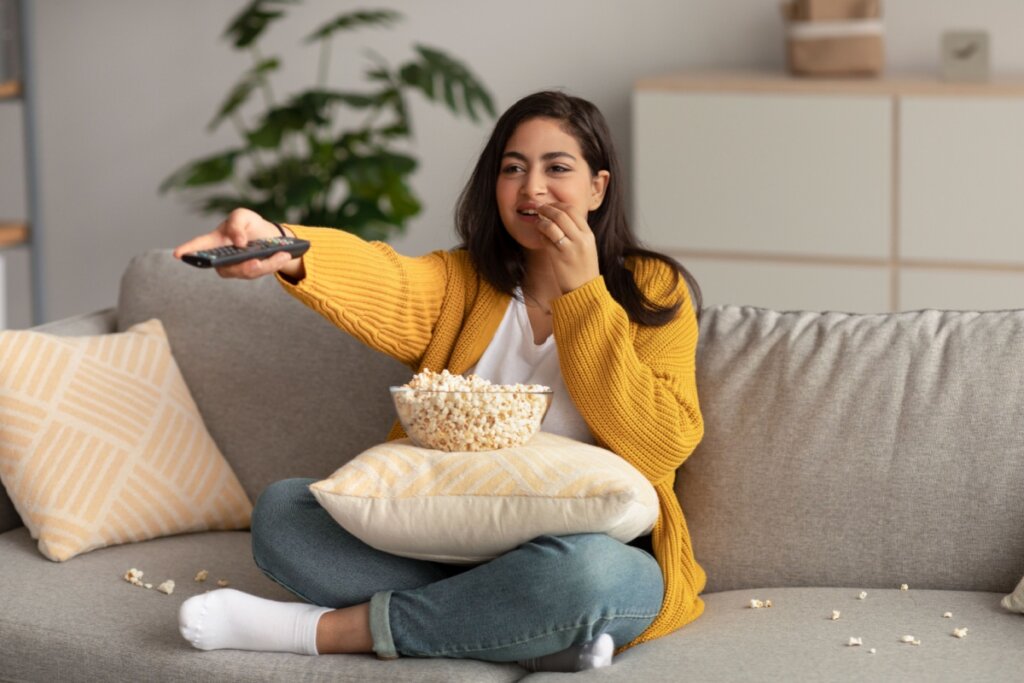 Mulher sentada no sofá comendo pipoca e segurando controle remoto de TV